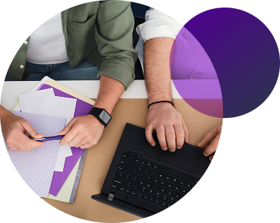 Aerial view of two people sitting at desk, with a notebook, pen, and laptop